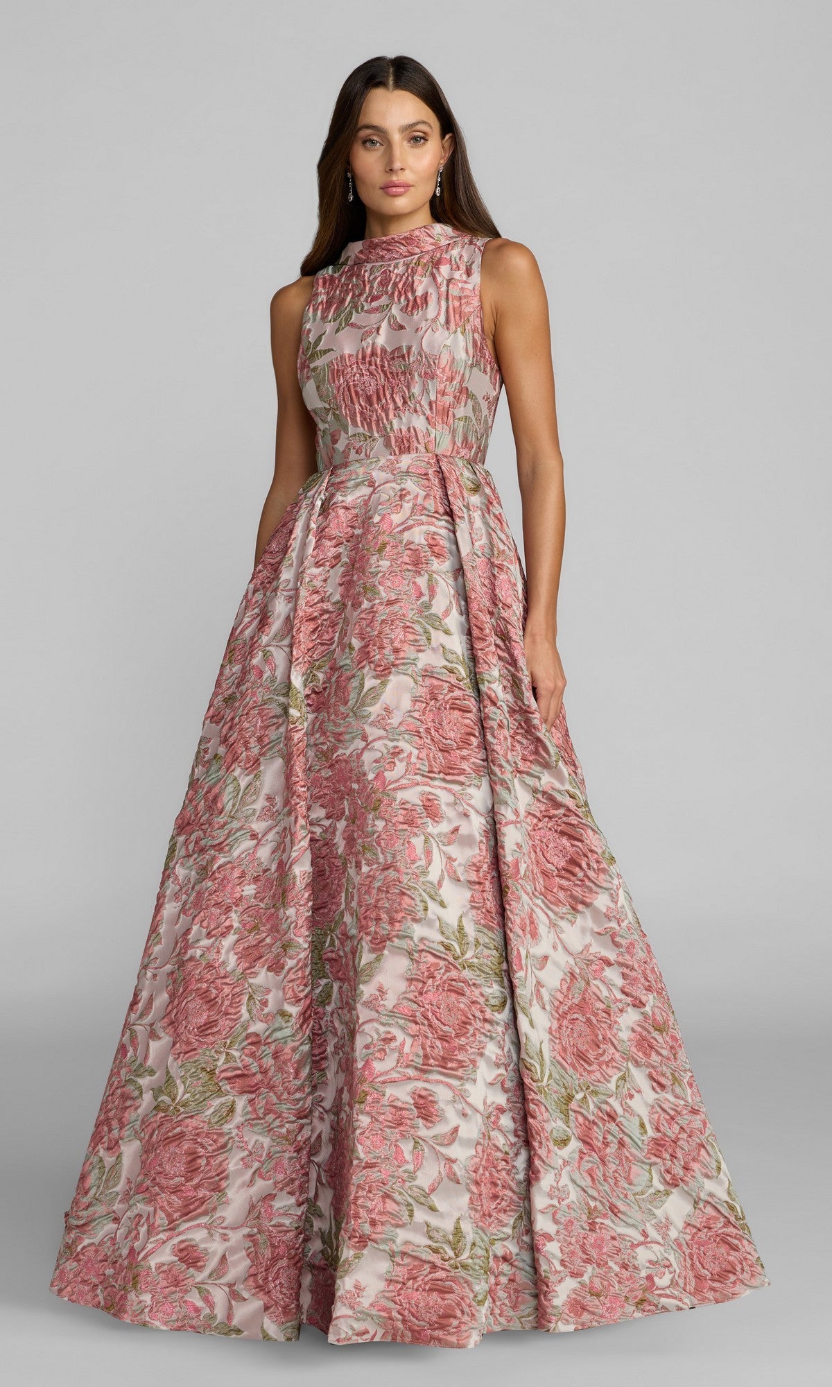 A woman wearing the Long Formal Dress: Mac Duggal 69177 in Rose Multi, featuring a sleeveless fitted bodice and a floor-length skirt with a beautiful pink floral brocade design, stands gracefully against a plain gray background.